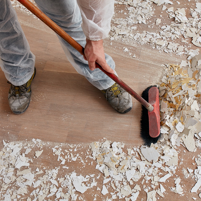 Ouvrier en combinaison blanche balayant des débris de plâtre cassés sur un sol en bois lors de travaux de rénovation.