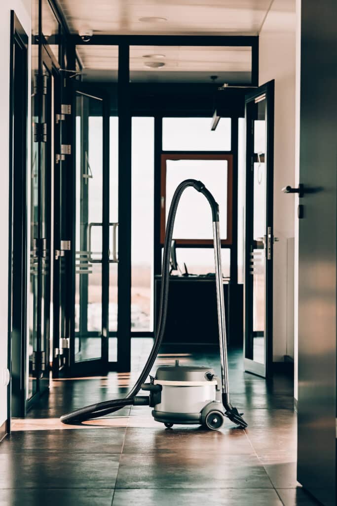 Aspirateur dans bureau moderne pour nettoyage professionnel Aspirateur traineau gris dans un couloir de bureau moderne avec portes vitrées ouvertes et lumière vive au fond.